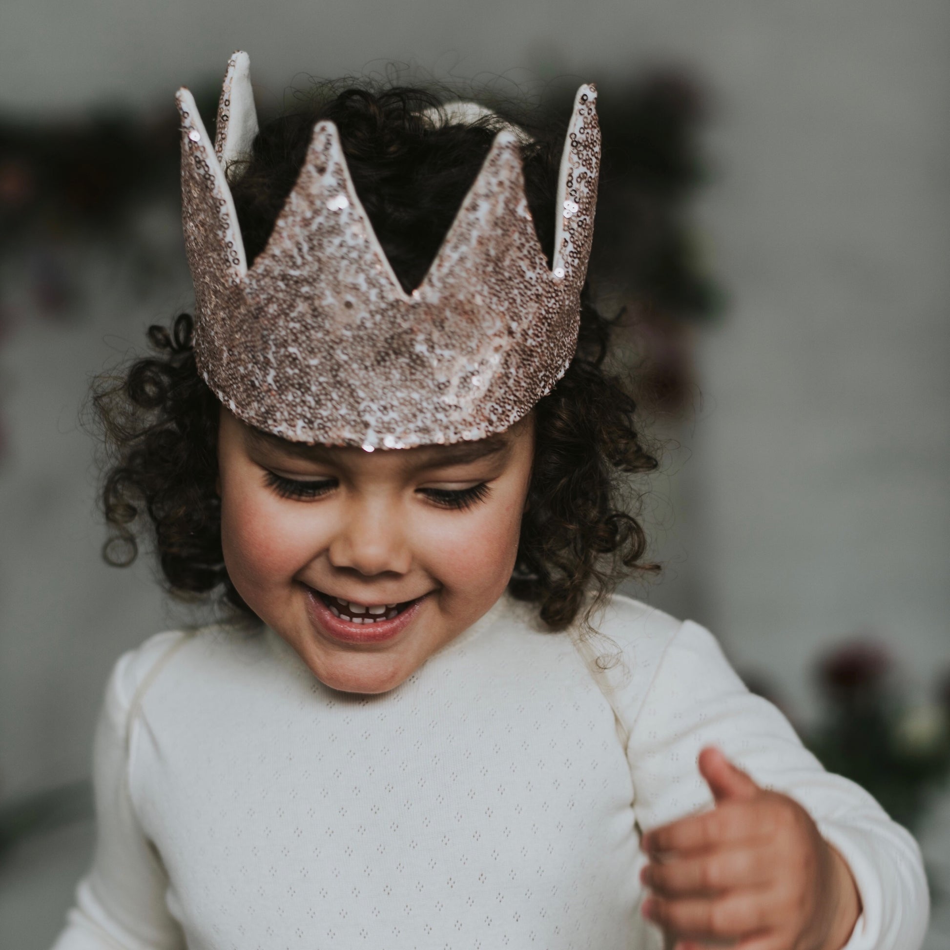Joyful child with curly hair wearing a handcrafted rose gold sequin crown, smiling during playtime.